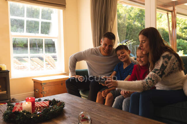 Famille caucasienne se détendre dans le salon à la période de Noël, assis sur le canapé, souriant et prendre selfie avec smartphone. temps de famille de qualité ensemble, célébration de Noël. — Photo de stock