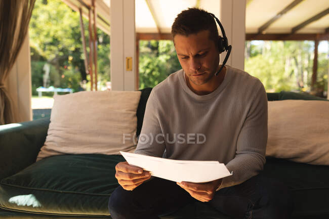 Caucasian man working from home wearing a phone headset and holding documents sitting in living room. self isolaton during covid 19 coronavirus pandemic. — Stock Photo