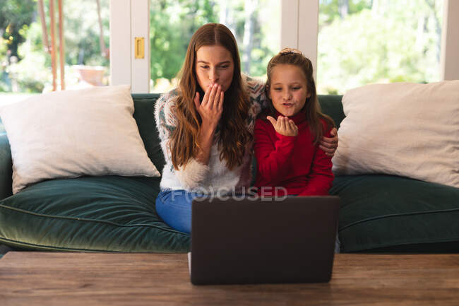 Mujer caucásica y su hija sentadas en el sofá y soplando besos, haciendo una videollamada usando un portátil. autoaislamiento durante el bloqueo de cuarentena del covid 19 del coronavirus. - foto de stock