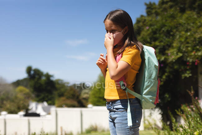 Chica caucásica con el pelo oscuro usando mascarilla caminando a la escuela llevando mochila. educación y estilo de vida durante la pandemia de coronavirus covid 19 - foto de stock
