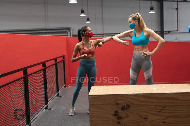 Caucasian female fitness trainer and fit caucasian woman wearing face masks greeting each other by touching elbows in the gym. social distancing quarantine lockdown during coronavirus pandemic — Stock Photo