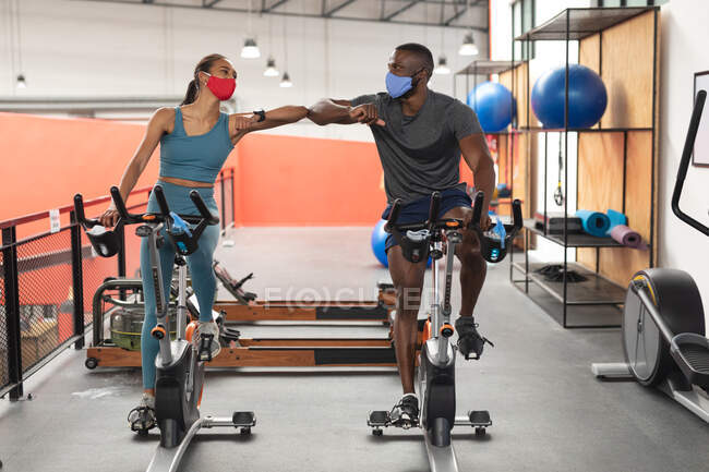 Fit african american man and fit caucasian woman wearing face masks greeting each other by touching elbows while exercising on stationary bike in the gym. social distancing quarantine lockdown during coronavirus pandemic — Stock Photo