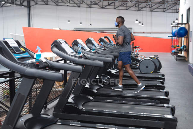 Fit afrikanisch-amerikanischer Mann mit Gesichtsmaske läuft auf dem Laufband beim Cardio-Training im Fitnessstudio. Soziale Distanzierung von Quarantäne während der Coronavirus-Pandemie — Stockfoto