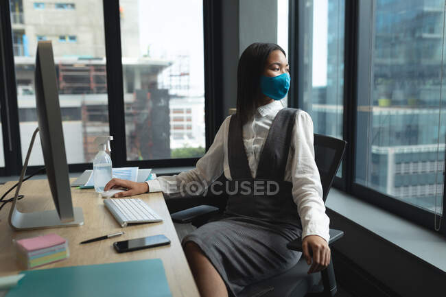 Mujer asiática con máscara facial sentada en su escritorio mirando por la ventana de la oficina moderna. distanciamiento social bloqueo de cuarentena durante la pandemia de coronavirus - foto de stock