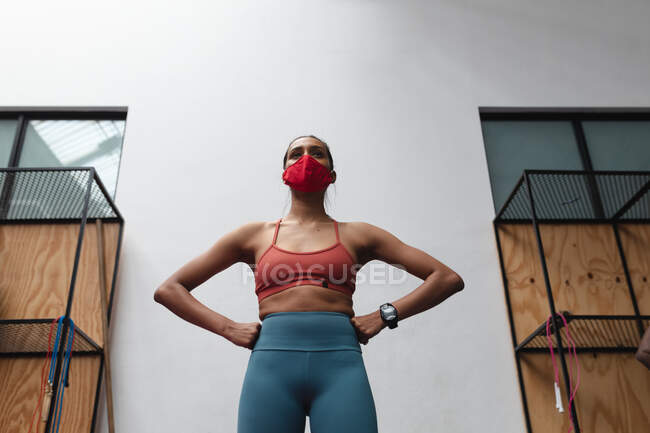 Fit femme caucasienne portant un masque facial debout avec la main sur sa taille dans la salle de gym. isolement social mise en quarantaine pendant une pandémie de coronavirus — Photo de stock