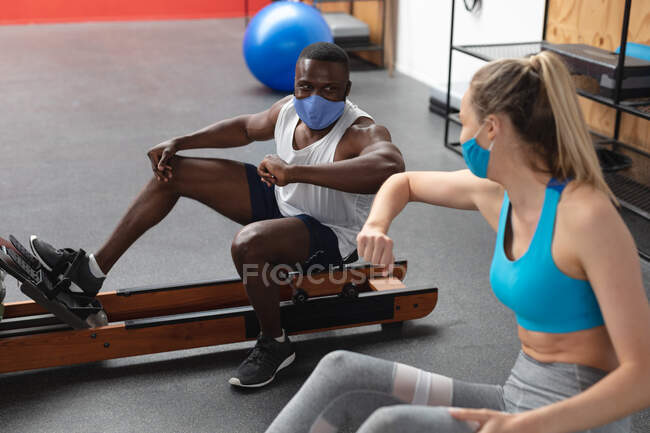 Fit african american man and fit  caucasian woman wearing face masks greeting each other by touching elbows while exercising in the gym. social distancing quarantine lockdown during coronavirus pandemic — Stock Photo