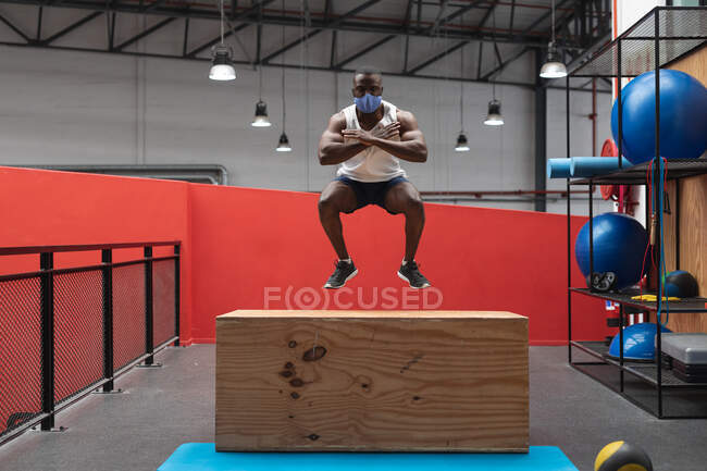 Жирний афроамериканець, одягнений в маску обличчя, стрибає на дерев'яній скриньці для слини під час тренування витривалості в спортзалі. Відстань до карантину під час пандемії коронавірусу — стокове фото