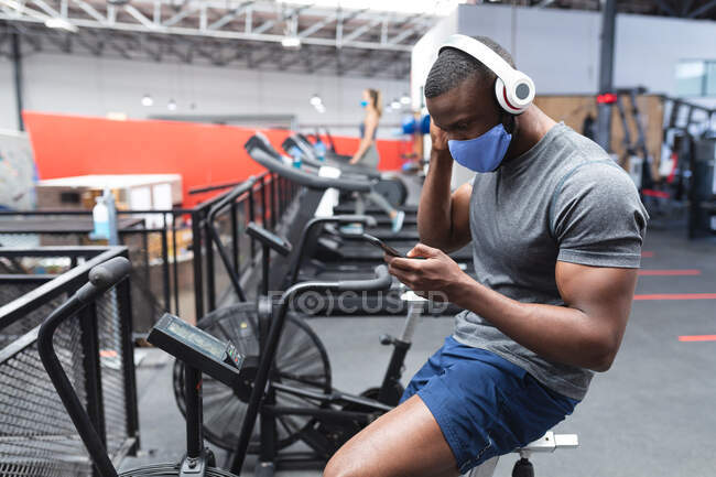 Жирний афроамериканець, одягнений в маску обличчя і навушники за допомогою смартфона, сидячи на стаціонарному велосипеді в спортзалі. Відстань до карантину під час пандемії коронавірусу — стокове фото