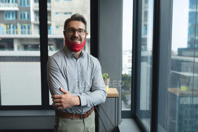 Portrait d'un homme caucasien avec un masque autour du cou, les bras croisés dans un bureau moderne. isolement social mise en quarantaine pendant une pandémie de coronavirus — Photo de stock