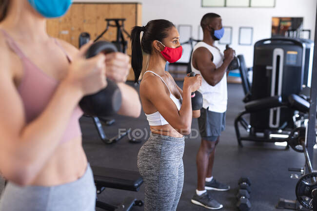 Fit african american man and two fit caucasian women wearing face masks performing exercise with kettlebells in the gym. social distancing quarantine lockdown during coronavirus pandemic — Stock Photo