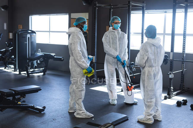 Equipo de trabajadores con ropa protectora discutiendo juntos en el gimnasio. distanciamiento social bloqueo de cuarentena durante la pandemia de coronavirus - foto de stock