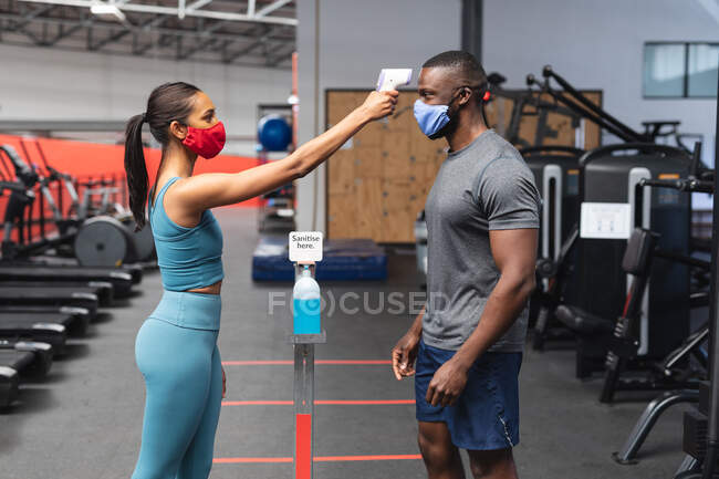 Fit caucasian woman wearing face mask checking temperature of fit african american man wearing face mask in the gym. social distancing quarantine lockdown during coronavirus pandemic — Stock Photo