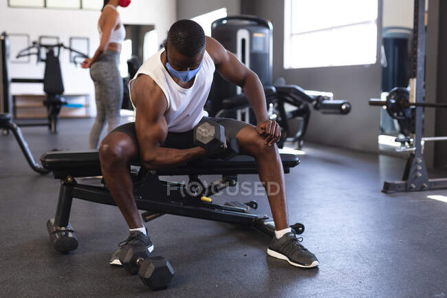 Convient à l'homme afro-américain portant un masque facial effectuant un exercice avec haltère dans la salle de gym. isolement social mise en quarantaine pendant une pandémie de coronavirus — Photo de stock