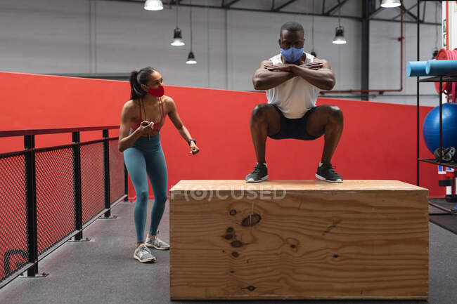 Ajuste hombre afroamericano con máscara facial saltando en la caja de madera del plyo en el gimnasio mientras que el entrenador de fitness femenino caucásico sostiene el cronómetro. distanciamiento social bloqueo de cuarentena durante la pandemia de coronavirus - foto de stock