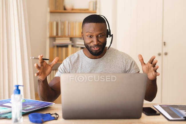 Африканский американец, использующий телефонные гарнитуры во время видеочата на ноутбуке, работая из дома. социальное дистанцирование во время блокады 19 коронавируса. — стоковое фото