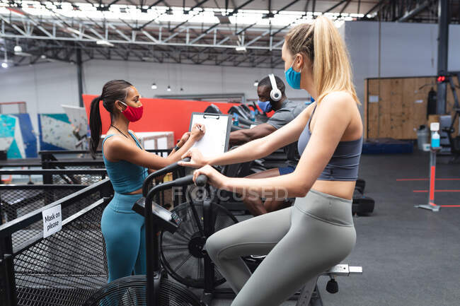 Fit caucasian woman wearing face mask exercising on stationary bike while caucasian female fitness coach taking notes on clipboard  in the gym. social distancing quarantine lockdown during coronavirus pandemic — Stock Photo