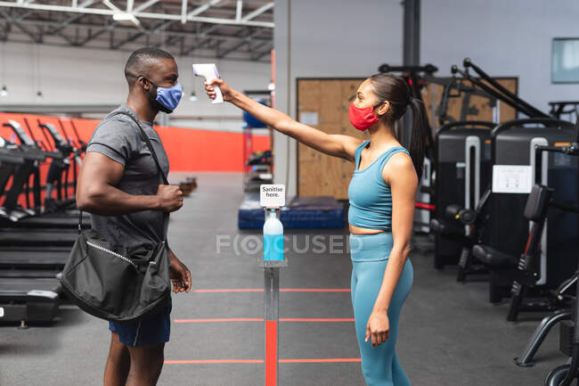 Fit caucasian woman wearing face mask checking temperature of fit african american man with gym bag in the gym. social distancing quarantine lockdown during coronavirus pandemic — Stock Photo