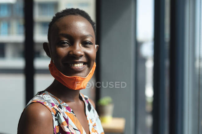 Portrait de femme afro-américaine avec un masque autour du cou souriant au bureau moderne. isolement social mise en quarantaine pendant une pandémie de coronavirus — Photo de stock