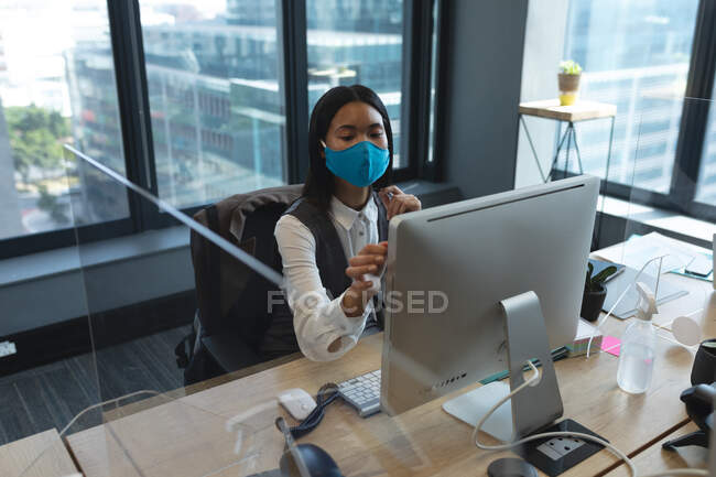 Mulher asiática usando máscara facial usando o computador enquanto se senta em sua mesa no escritório moderno. bloqueio de quarentena por distanciamento social durante a pandemia do coronavírus — Fotografia de Stock