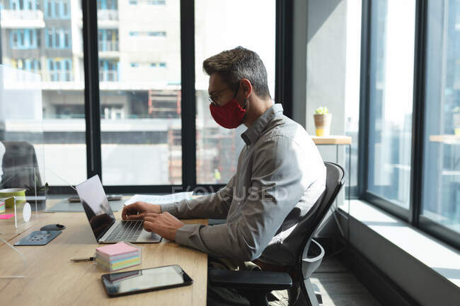 Белый мужчина в маске с ноутбуком, сидя на столе в современном офисе. социальная изоляция от карантина во время пандемии коронавируса — стоковое фото