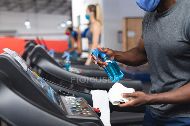Mid section of fit african american man wearing face mask sanitizing cardio machine before working out in the gym. social distancing quarantine lockdown during coronavirus pandemic — Stock Photo