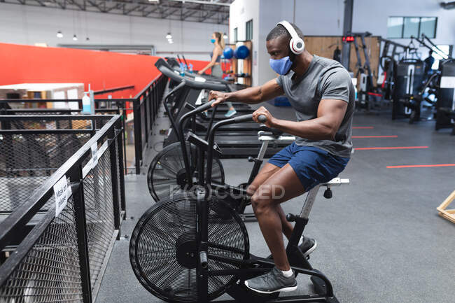 Жирний афроамериканець, одягнений в маску обличчя і навушники, працює на стаціонарному велосипеді в спортзалі. Відстань до карантину під час пандемії коронавірусу — стокове фото