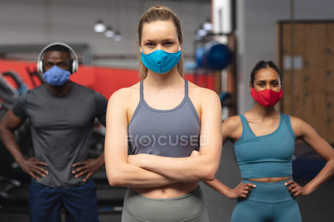 Portrait d'un homme afro-américain en forme et de deux femmes caucasiennes en forme portant des masques faciaux debout dans la salle de gym. isolement social mise en quarantaine pendant une pandémie de coronavirus — Photo de stock