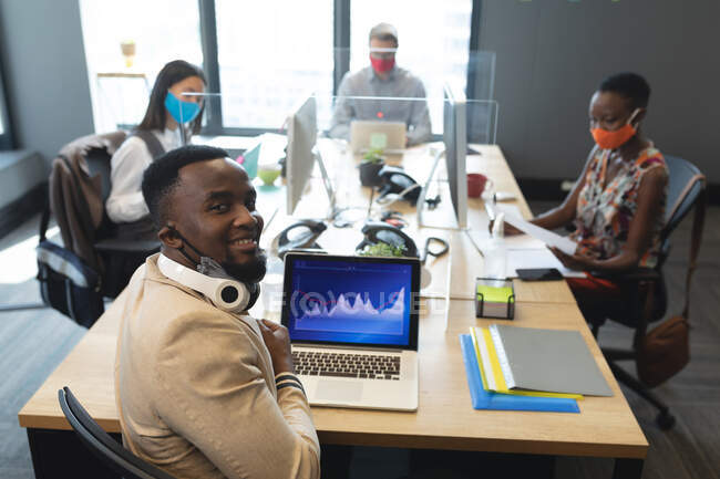 Retrato del hombre afroamericano usando auriculares usando laptop mientras está sentado en su escritorio en la oficina moderna. distanciamiento social bloqueo de cuarentena durante la pandemia de coronavirus - foto de stock