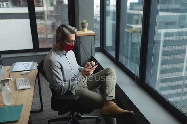 Белый мужчина в маске с помощью смартфона сидит на столе в современном офисе. социальная изоляция от карантина во время пандемии коронавируса — стоковое фото