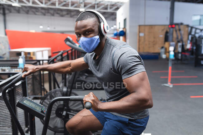 Portrait d'un homme afro-américain en forme portant un masque facial et des écouteurs faisant de l'exercice sur un vélo d'appartement dans la salle de gym. isolement social mise en quarantaine pendant une pandémie de coronavirus — Photo de stock