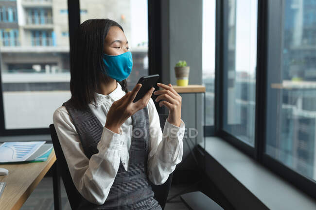 Жінка в азіатській масці говорить на смартфоні в сучасному офісі. Відстань до карантину під час пандемії коронавірусу — стокове фото