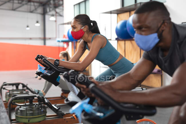 Fit afrikanisch-amerikanischer Mann und fit-kaukasische Frau mit Gesichtsmasken, die auf stationären Fahrrädern in der Turnhalle trainieren. Soziale Distanzierung von Quarantäne während der Coronavirus-Pandemie — Stockfoto