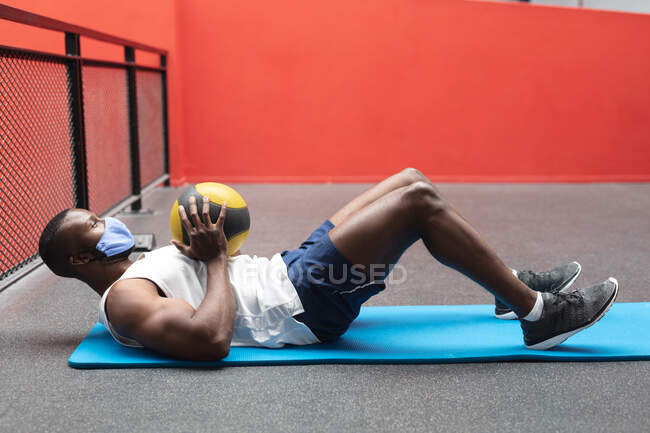 Подходящий африканский американец в маске для лица, выполняющий упражнения с мячом в спортзале. социальная изоляция от карантина во время пандемии коронавируса — стоковое фото