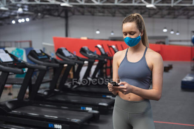 Ajuste mujer caucásica con mascarilla usando teléfono inteligente en el gimnasio. distanciamiento social bloqueo de cuarentena durante la pandemia de coronavirus - foto de stock