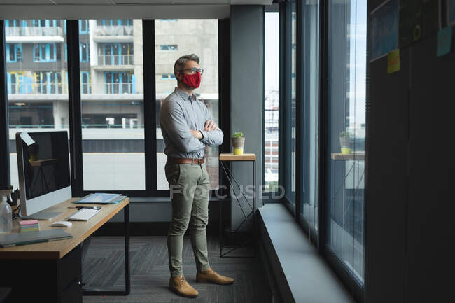 Caucasian man wearing face mask with arms crossed looking out of window at modern office. social distancing quarantine lockdown during coronavirus pandemic — Stock Photo