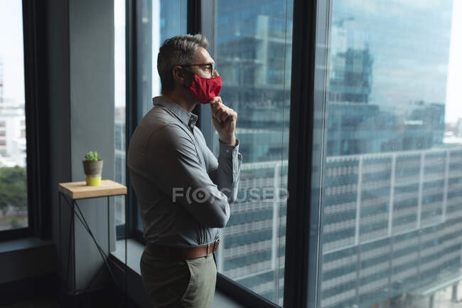 Un uomo caucasico premuroso che indossa una maschera che guarda fuori dalla finestra in un ufficio moderno. isolamento di quarantena a distanza sociale durante la pandemia di coronavirus — Foto stock