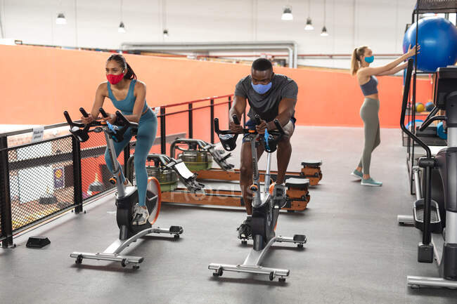 Fit african american man and fit caucasian woman wearing face masks exercising on stationary bike in the gym. social distancing quarantine lockdown during coronavirus pandemic — Stock Photo