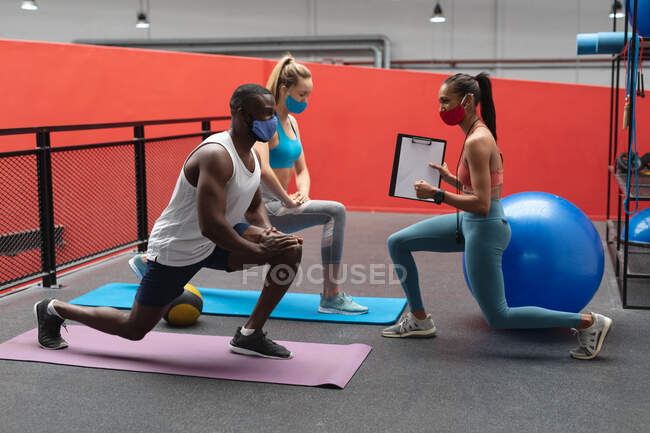 Entraîneur de fitness femme caucasienne tenant chronomètre et presse-papiers instruisant homme afro-américain en forme et femme caucasienne en forme pour effectuer des exercices d'étirement dans la salle de gym. — Photo de stock