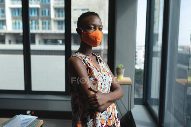 Портрет афро-американської жінки, одягненої в маску обличчя зі схрещеними руками в сучасному офісі. Відстань до карантину під час пандемії коронавірусу — стокове фото