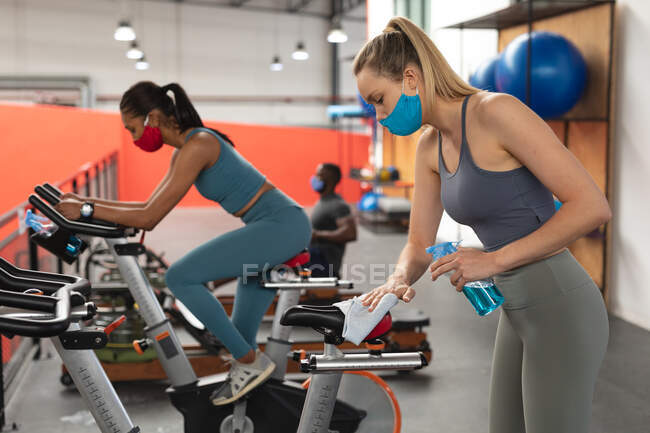 Fit caucasian woman wearing face mask sanitizing seat of stationary bike before working out in the gym. social distancing quarantine lockdown during coronavirus pandemic — Stock Photo