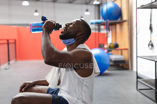 Un homme afro-américain avec un masque autour du cou qui boit de l'eau au gymnase. isolement social mise en quarantaine pendant une pandémie de coronavirus — Photo de stock
