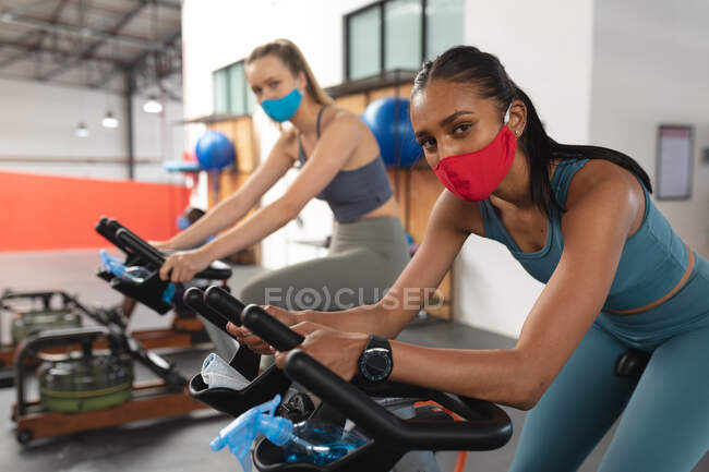 Retrato de duas mulheres caucasianas em forma usando máscaras faciais exercitando-se em bicicleta estacionária no ginásio. bloqueio de quarentena por distanciamento social durante a pandemia do coronavírus — Fotografia de Stock
