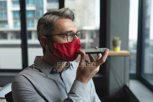 Kaukasischer Mann mit Gesichtsmaske, der im modernen Büro mit dem Smartphone spricht. Soziale Distanzierung von Quarantäne während der Coronavirus-Pandemie — Stockfoto