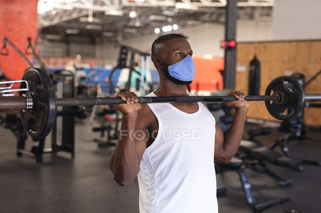 Жирний афроамериканець, одягнений у маску обличчя, виконує вправи з дзвінком у спортзалі. Відстань до карантину під час пандемії коронавірусу — стокове фото