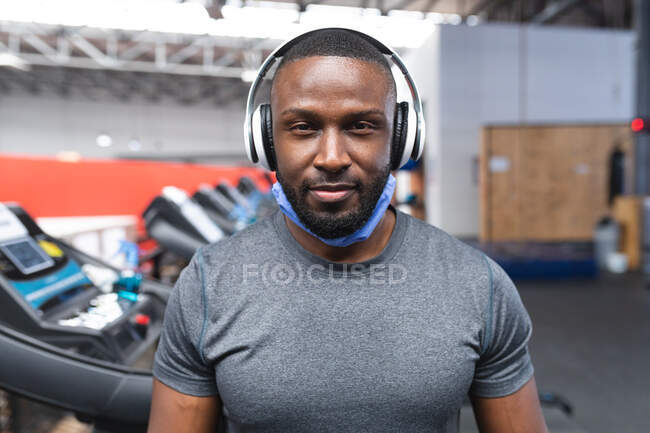 Portrait d'un homme afro-américain en forme avec un masque autour du cou portant des écouteurs dans la salle de gym. isolement social mise en quarantaine pendant une pandémie de coronavirus — Photo de stock