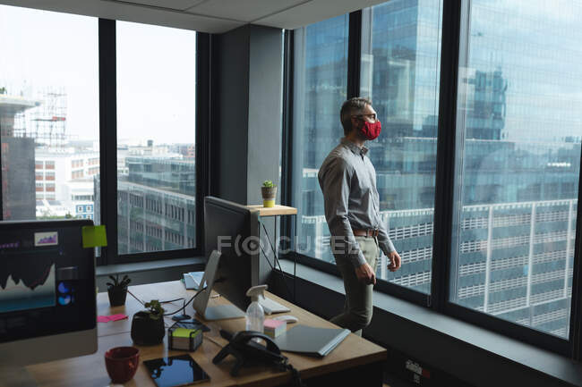 Uomo caucasico che indossa maschera viso guardando fuori dalla finestra in ufficio moderno. isolamento di quarantena a distanza sociale durante la pandemia di coronavirus — Foto stock