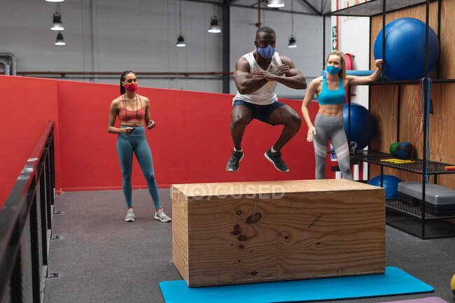 Подходящий африканский американец в маске для лица, прыгающий на деревянной пластиковой коробке в спортзале, в то время как белая женщина-тренер по фитнесу держит секундомер. социальная изоляция от карантина во время пандемии коронавируса — стоковое фото
