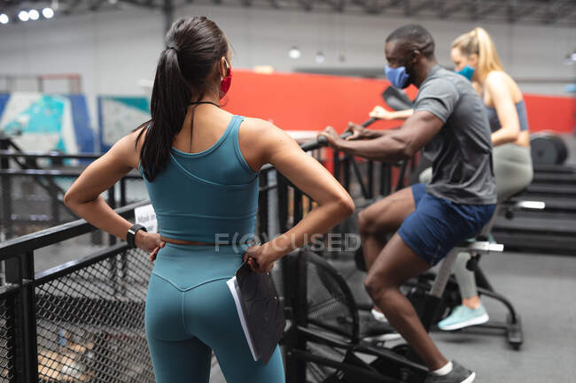 Fit african american man wearing face mask exercising on stationary bike while caucasian female fitness coach with clipboard inspecting in the gym. social distancing quarantine lockdown during coronavirus pandemic — Stock Photo