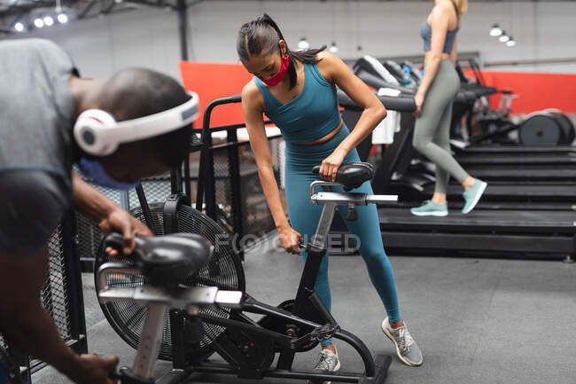 Жирний афро-американський чоловік і підходяща жінка з кавказькими масками, що регулюють місце стаціонарного велосипеда в спортзалі. Відстань до карантину під час пандемії коронавірусу — стокове фото