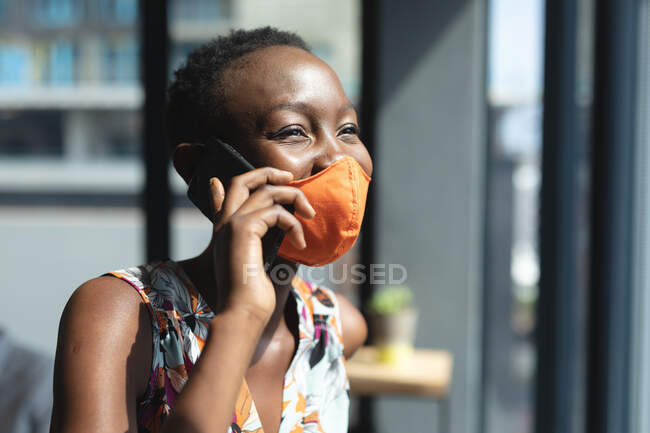 Femme afro-américaine portant un masque facial parlant sur smartphone au bureau moderne. isolement social mise en quarantaine pendant une pandémie de coronavirus — Photo de stock